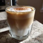 Zbliżenie na szklankę caffe latte macchiato przepis z warstwami mleka, kawy i puszystej pianki posypanej cynamonem.