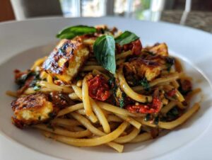 Zbliżenie na makaron z halloumi i suszonymi pomidorami, udekorowany świeżą bazylią, podany na białym talerzu.
