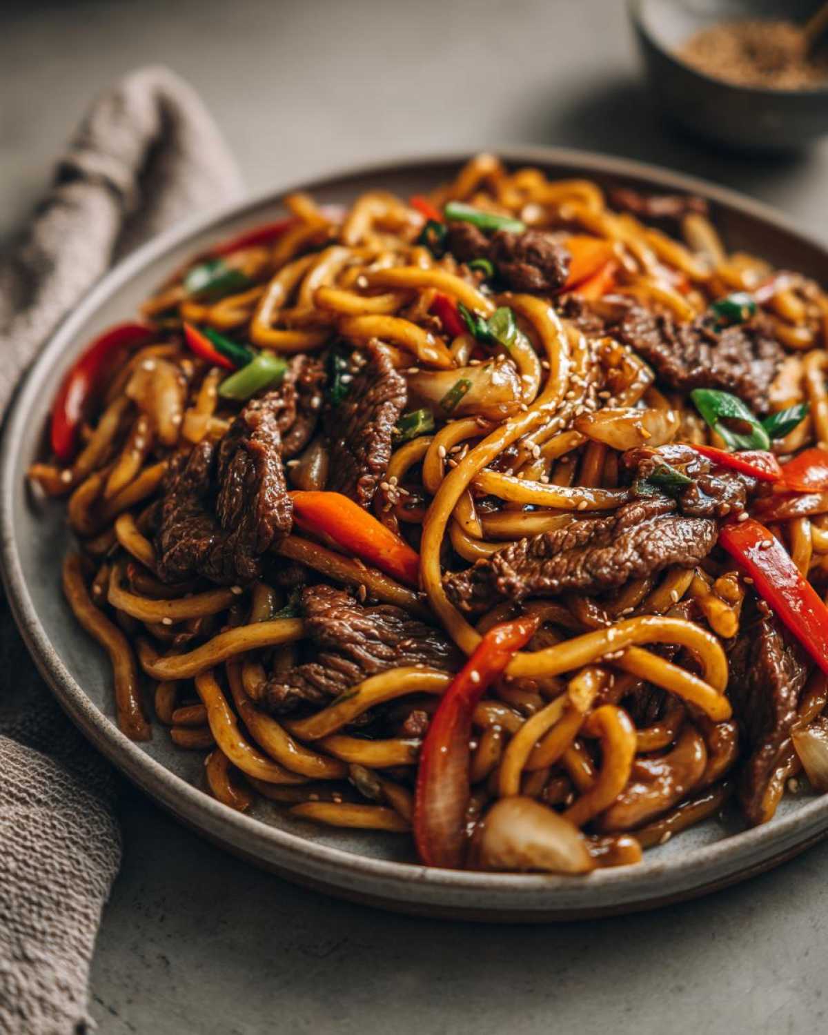 Zbliżenie na danie makaron udon z wołowiną, papryką, cebulą i sezamem na szarym talerzu.