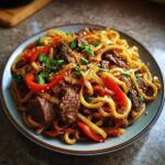 Zbliżenie na talerz z makaronem udon z wołowiną, czerwoną papryką, cebulą i szczypiorkiem, posypany sezamem.