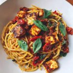 Zbliżenie na makaron z halloumi i suszonymi pomidorami, udekorowany świeżymi liśćmi bazylii w białej misce.