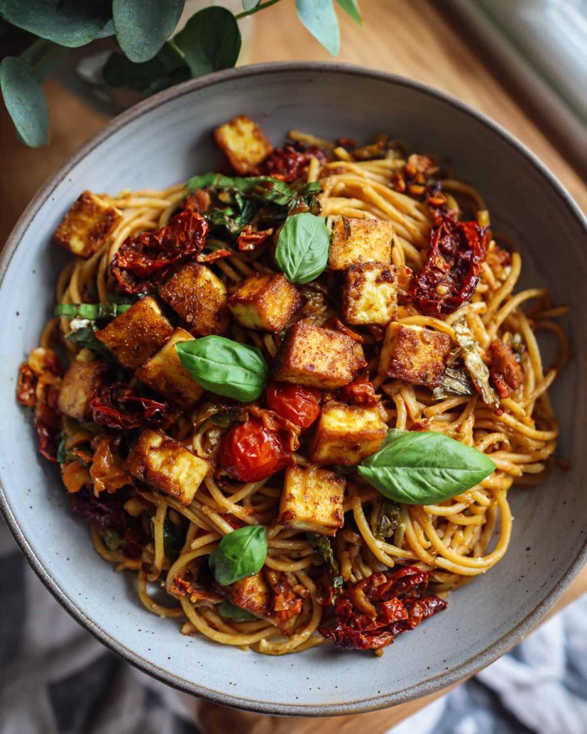 Zbliżenie miski makaronu z halloumi i suszonymi pomidorami, udekorowanego świeżymi liśćmi bazylii.
