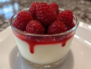 Zbliżenie na szklankę panna cotta oryginalny przepis z sosem malinowym i świeżymi malinami na wierzchu.