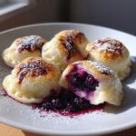 Zbliżenie na talerz z kilkoma pierogami z jagodami, posypanymi cukrem pudrem, jeden jest przekrojony, ukazując soczyste nadzienie jagodowe. pierogi z jagodami.
