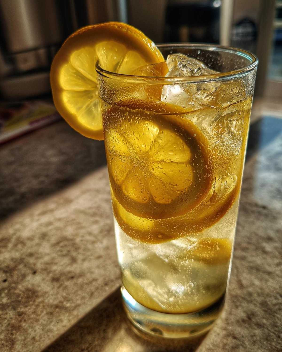 Zbliżenie na szklankę lemoniady z plasterkami cytryny i kostkami lodu, stojącą w słońcu.