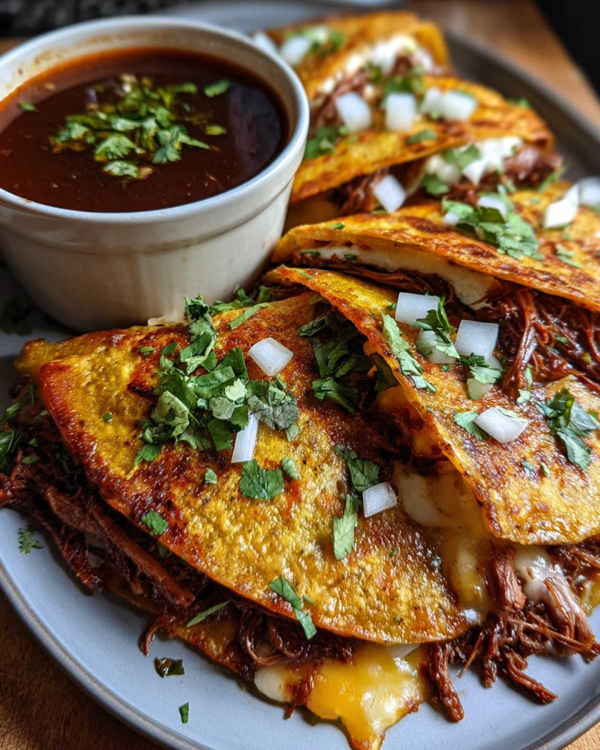 Zbliżenie na talerz z tacos z wołowiną birria, serem i posiekaną cebulą, podane z miseczką consommé.