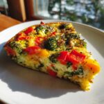 Kawałek frittata warzywna z brokułami, czerwoną i żółtą papryką na talerzu.