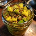 Słoik pełen kawałków kiszonego mango z przyprawami, w tym gorczycą i liściem laurowym.