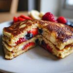 Zbliżenie na stos naleśników pełnoziarnistych fit z truskawkami i jagodami w środku, podane z owocami.