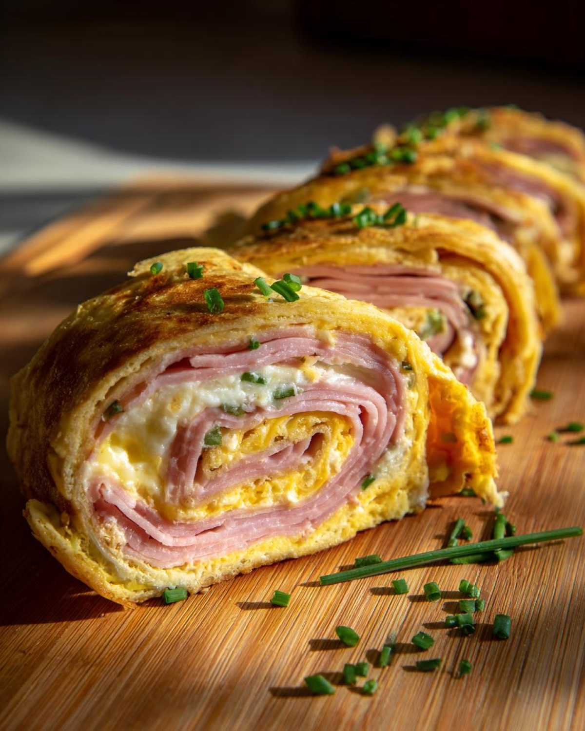 Krojone rollsy z tortilli z szynką, jajkiem i serem, posypane szczypiorkiem na drewnianej desce.