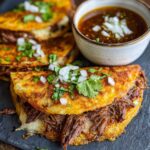 Zbliżenie na tacos z wołowiną birria, ser i kolendrę, obok miseczka z bulionem.