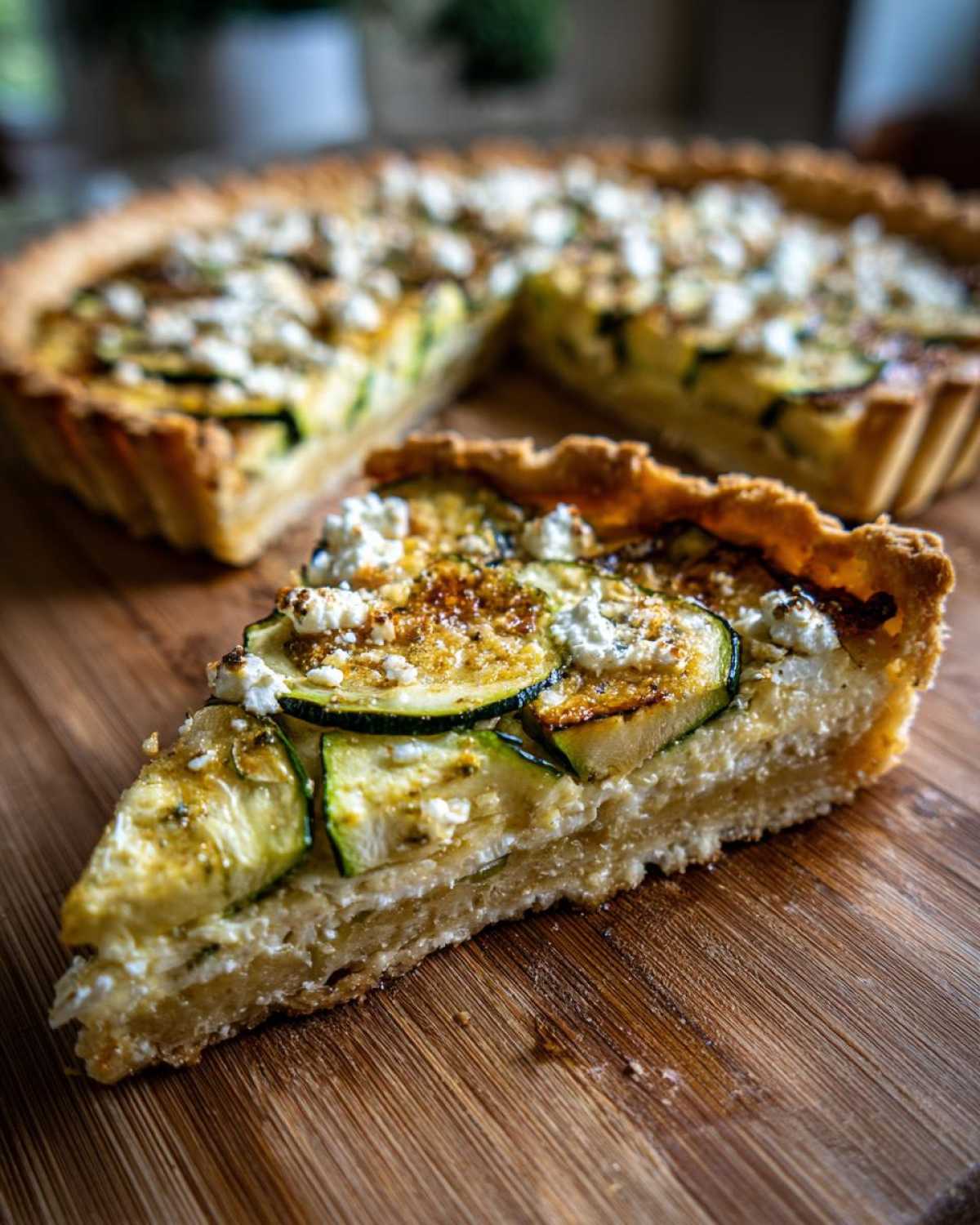 Zdjęcie przedstawia kawałek tarta z cukinią i fetą na drewnianej desce. Widoczna cała tarta w tle.