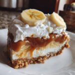 Kawałek pysznego banoffee pie bez pieczenia z bananami, karmelem i bitą śmietaną na białym talerzu.
