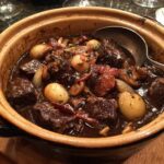 Zbliżenie na Boeuf Bourguignon w glinianym naczyniu, z widocznymi kawałkami wołowiny, cebulą i grzybami.