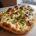 Zbliżenie na puszysty chlebek naan czosnkowy posypany posiekaną natką pietruszki i polany masłem.
