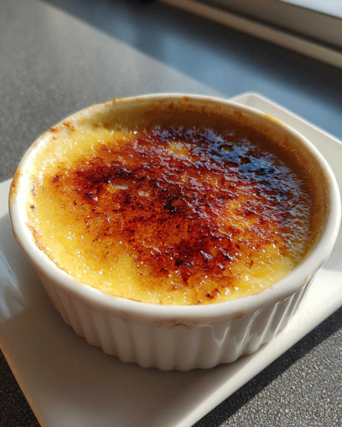 Zbliżenie na deser Crème brûlée w białej kokilce z idealnie skarmelizowaną, brązową warstwą cukru.
