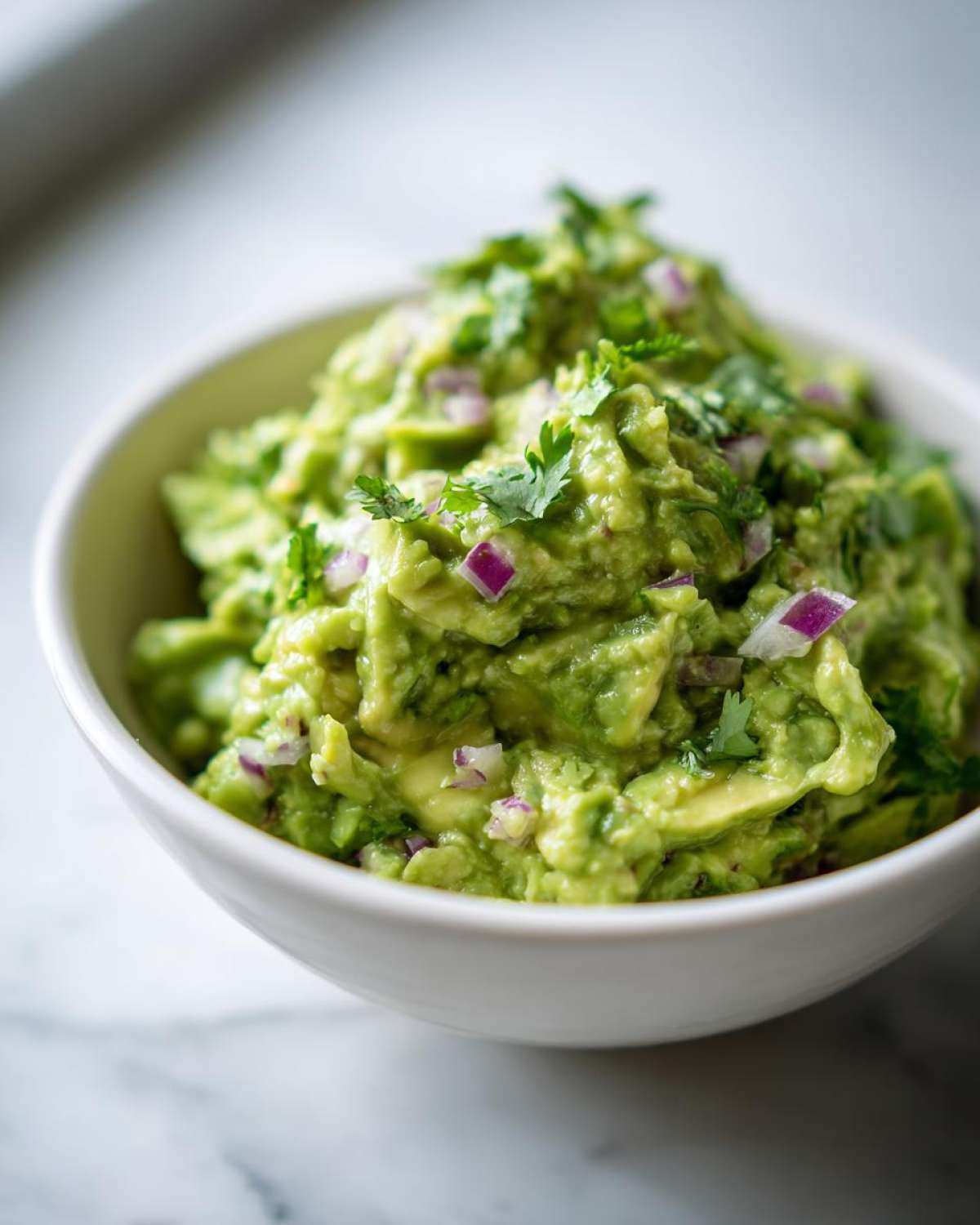 Zbliżenie na miskę z prostym guacamole domowym, ozdobionym posiekaną kolendrą i czerwona cebulą.
