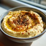 Aksamitny klasyczny hummus w misce, polany oliwą i posypany przyprawami i sezamem.