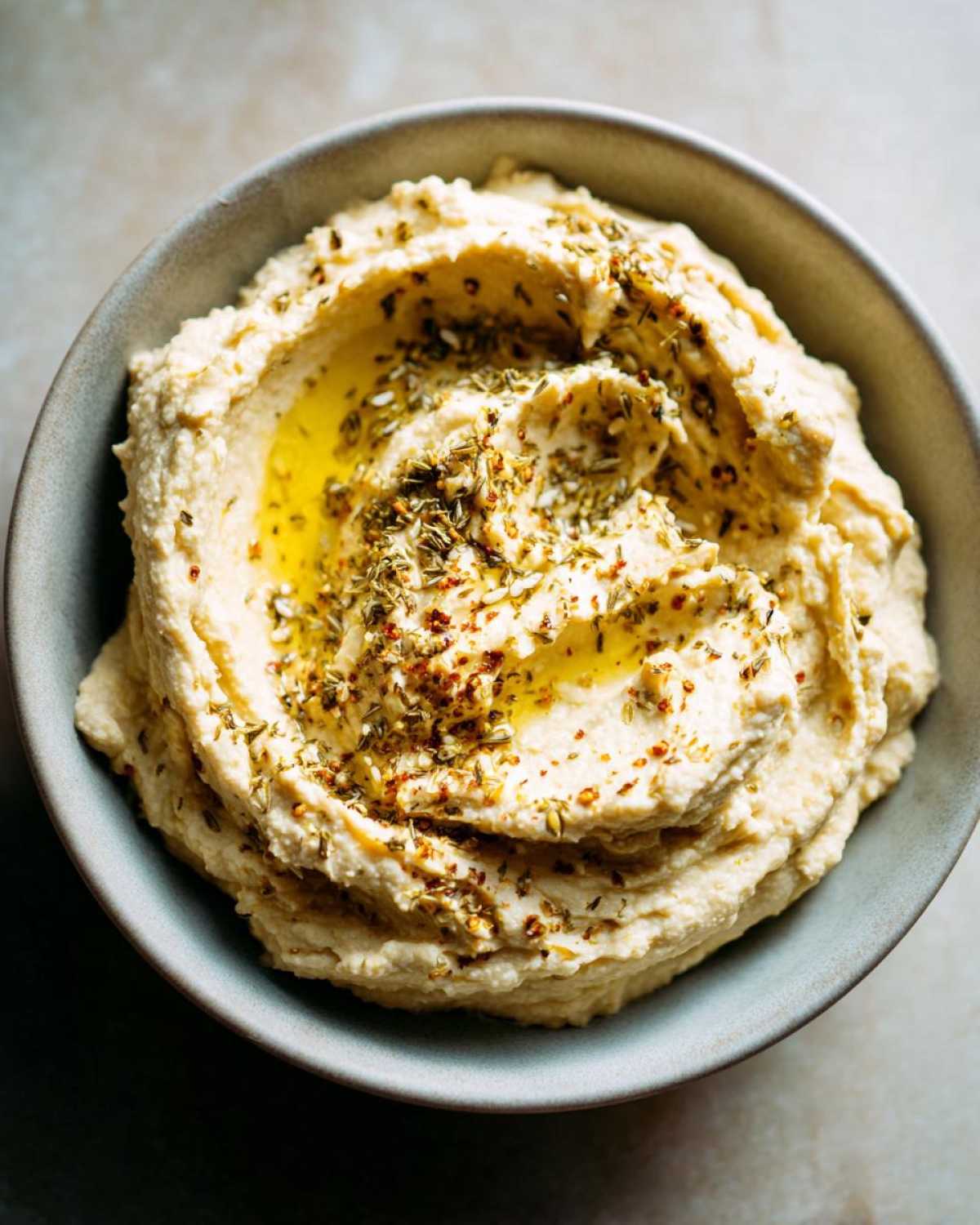 Aksamitny klasyczny hummus w misce, polany oliwą i posypany ziołami i papryką.