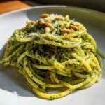 Zbliżenie na talerz makaronu z pesto bazyliowym, posypany parmezanem i orzeszkami pinii.