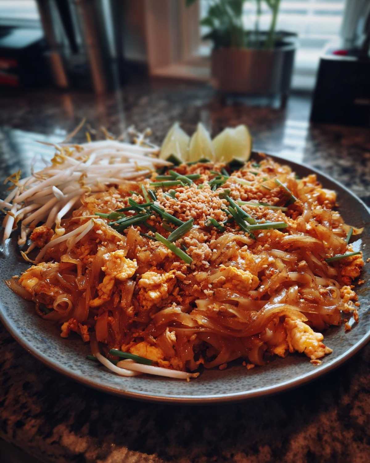 Talerz pełen Pad Thai Klasyczny, ozdobiony szczypiorkiem, orzeszkami ziemnymi i ćwiartkami limonki.