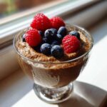Pudding chia czekoladowy bez cukru udekorowany świeżymi malinami i borówkami w szklanej miseczce.