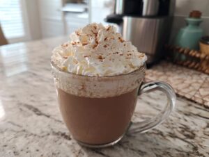 Szklana filiżanka z Pumpkin Spice Latte, ozdobiona obfitą porcją bitej śmietany i posypką z cynamonu.