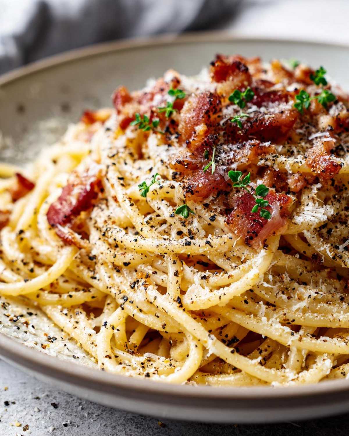 Zbliżenie na talerz spaghetti carbonara z chrupiącym boczkiem, startym parmezanem i świeżym pieprzem.