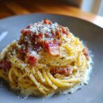 Zbliżenie na talerz z porcją spaghetti carbonara, udekorowane kawałkami boczku, startym parmezanem i świeżo mielonym pieprzem.