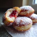 Zbliżenie na pączki domowe posypane cukrem pudrem, jeden pączek przekrojony ukazuje wnętrze z dżemem.