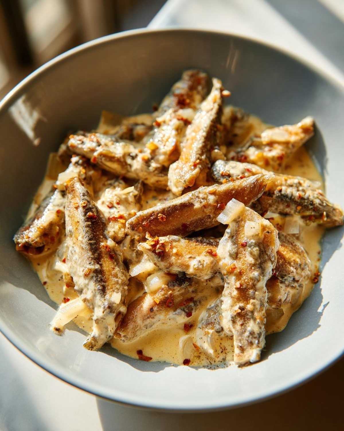Zbliżenie na miskę pełną śledzi w śmietanie, posypanych płatkami chili i cebulą.