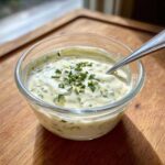 Zbliżenie na pyszny sos czosnkowy w szklanej miseczce, posypany ziołami, z łyżeczką w środku.