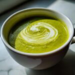 Zbliżenie na miskę zupy krem z brokułów z wirującą śmietaną na wierzchu, ozdobioną ziołami.