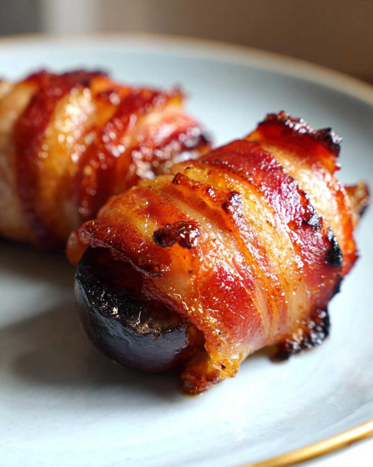Zbliżenie na dwa kawałki boczku ze śliwką, zawinięte w chrupiący boczek, podane na talerzu.