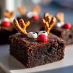 Zbliżenie na brownie reniferki z oczami z masy cukrowej i nosem z czerwonej drażetki, ozdobione precelkami jako rogami.