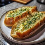 Dwa kawałki chleba czosnkowego posypane posiekaną pietruszką, leżące na białym talerzu.