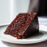 Kawałek wilgotnego, ciemnego ciasto czekoladowe z chilli, widoczne czerwone drobinki przyprawy, na jasnym talerzyku.