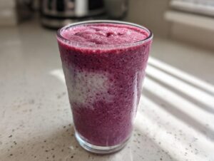 Szklanka z gęstym, fioletowym shake jagodowym z widocznymi warstwami, stojąca na jasnym blacie.