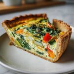 Zbliżenie na kawałek quiche z warzywami, szpinakiem i papryką na białym talerzu.