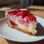 Zbliżenie na kawałek kremowego sernika truskawkowego z warstwą owoców na wierzchu, podany na białym talerzu.