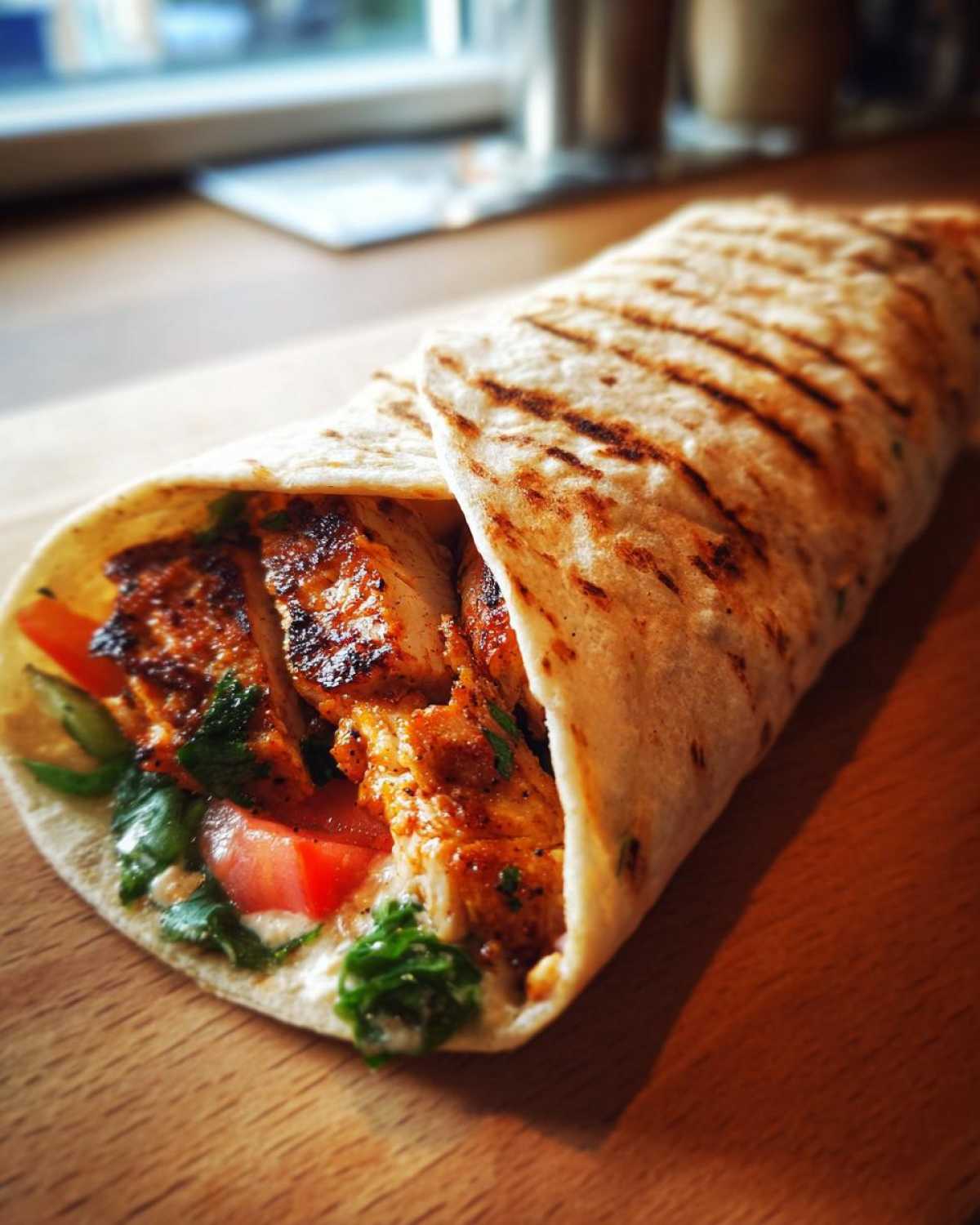 Zbliżenie na domowy kebab z kurczaka w grillowanej tortilli, z kawałkami kurczaka, pomidorem i zielonymi ziołami.