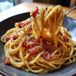 Porcja kremowej pasta carbonara z kawałkami boczku i dużą ilością świeżo mielonego pieprzu, podnoszona widelcem.