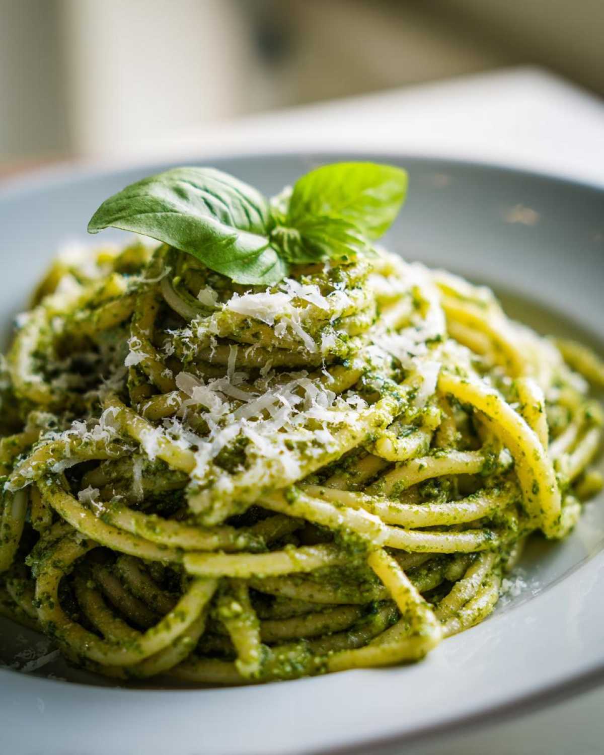 Zbliżenie na talerz z makaronem z pesto, posypanym parmezanem i listkiem bazylii.