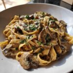 Zbliżenie na talerz pełen makaronu tagliatelle z kremowym makaron z sosem grzybowym i posiekaną natką pietruszki.