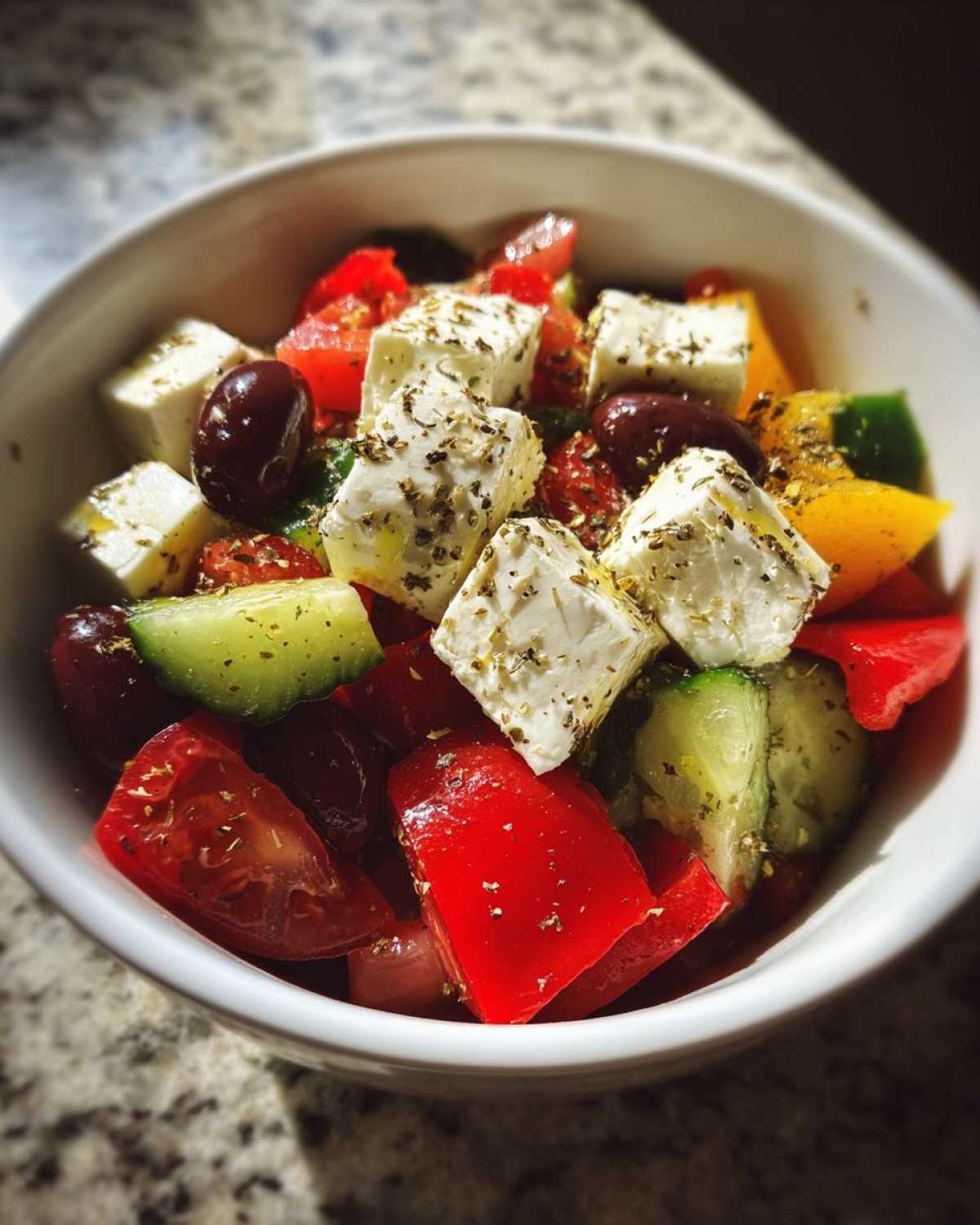 Zbliżenie na miskę pełną sałatki greckiej z serem feta, pomidorami, ogórkiem i oliwkami, posypanej ziołami.