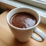 Zbliżenie na gęstą, aksamitną Gorąca czekolada domowa w białym kubku, stojącym na drewnianym blacie.