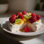 Przekrojona Beza Pavlova z bitą śmietaną, malinami, kiwi i musem z marakui na białym talerzu.