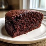 Zbliżenie na kawałek wilgotnego ciasta czekoladowego o intensywnie brązowym kolorze na białym talerzyku.
