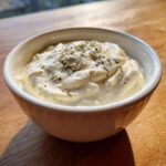 Kremowy dip czosnkowy domowy posypany suszonymi ziołami w białej miseczce na drewnianym stole.