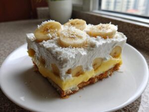 Duży kawałek ciasta Bananowa chmurka z warstwą budyniu, bitej śmietany i plasterkami banana.